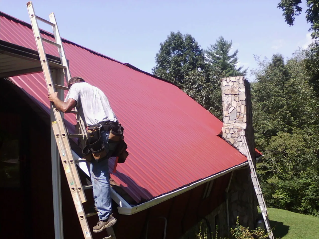 Roofer on a ladder beside a red metal roof and stone chimney — Soffit & Fascia Repair by Roofing Brothers in Mount Airy
